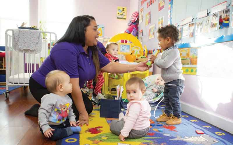 Classroom with babies and toddlers. Teacher holds a toy, and a toddler boy pretends it is a microphone.