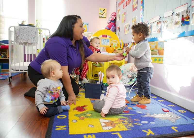Classroom with babies and toddlers. Teacher holds a toy, and a toddler boy pretends it is a microphone.