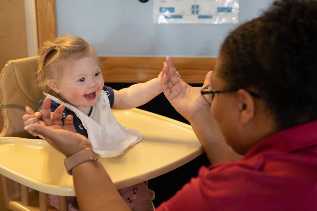 Child in high-chair smiles and looks at educator as they hold hands