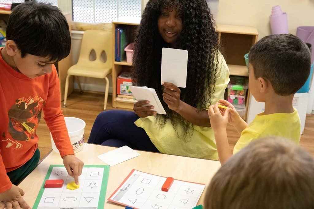 Educator sits with three children at table. They hold up a card. One child points to a hexagon. Another child holds a shape.
